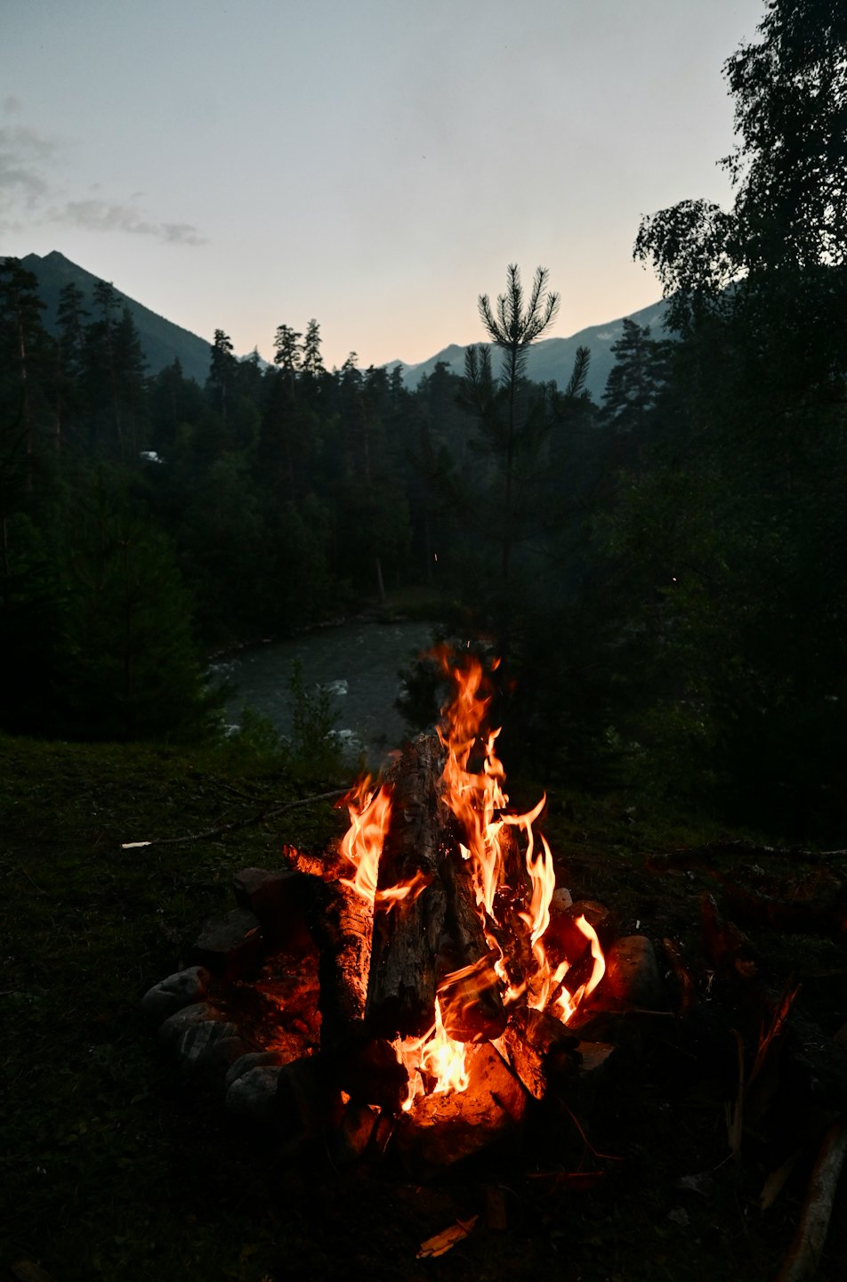 A campfire burns inside a stone pit near a river.